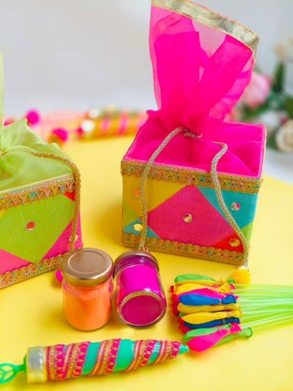 Colorful Holi basket hamper with two zars of colors, a pichkari, and a balloon bunch.