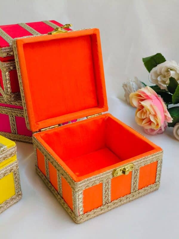 An orange hamper box with a textured surface, featuring a hinged lid and a matching ribbon tie. The box is placed on a wooden table, with soft lighting that highlights its vibrant color and elegant design