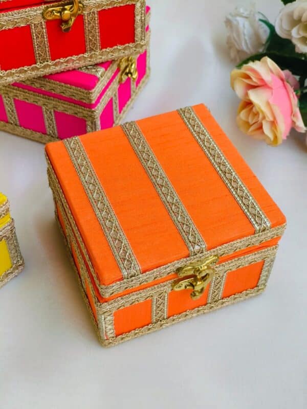 An orange hamper box with a textured surface, featuring a hinged lid and a matching ribbon tie. The box is placed on a wooden table, with soft lighting that highlights its vibrant color and elegant design